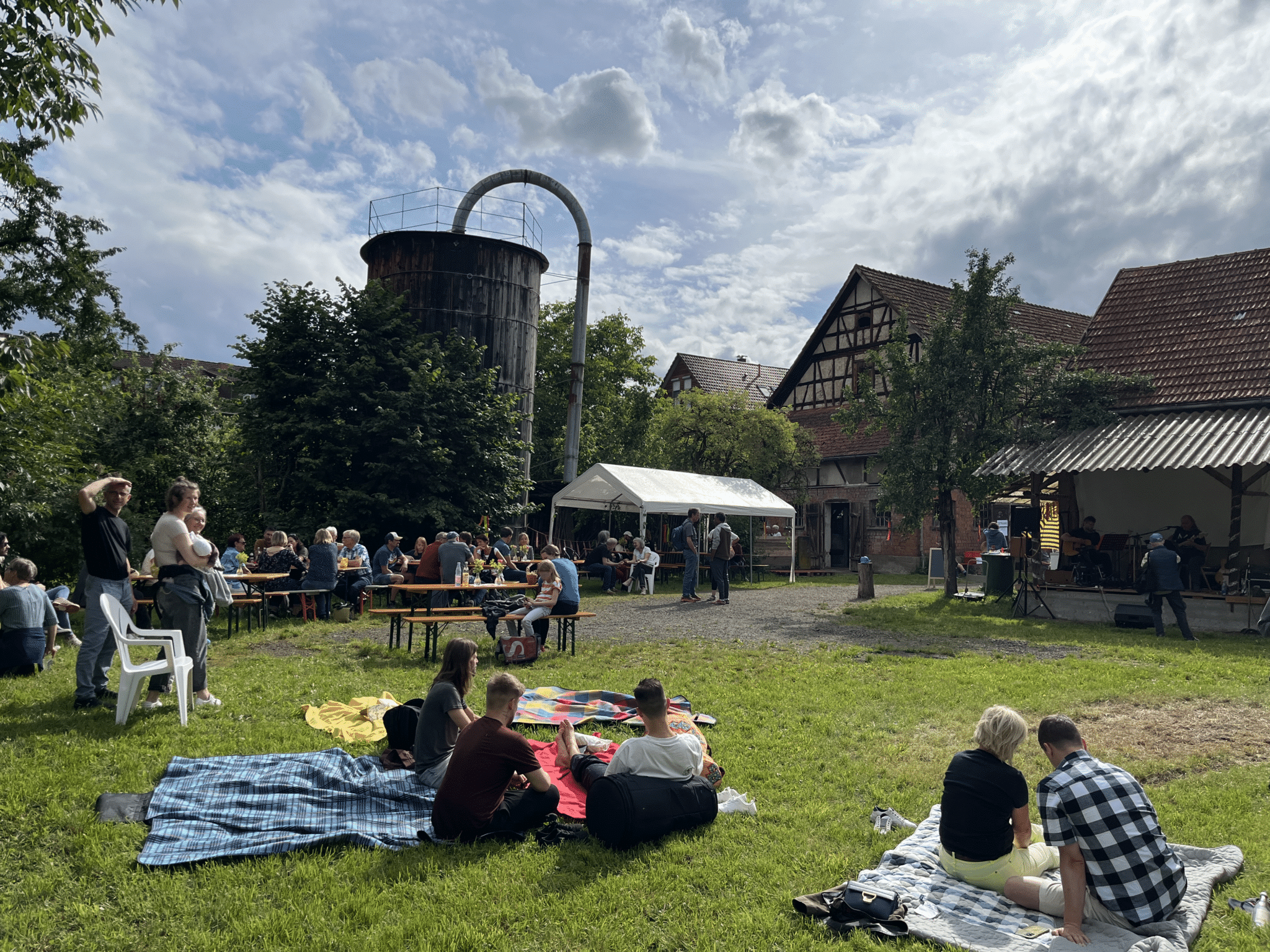 Menschen sitzen auf Picknickdecken, im Hintergrund alte Bauernhof-Gebäude.